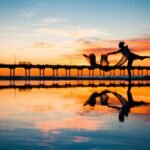 Silhouette of a dancer performing near a jetty at sunset with vibrant sky reflection.